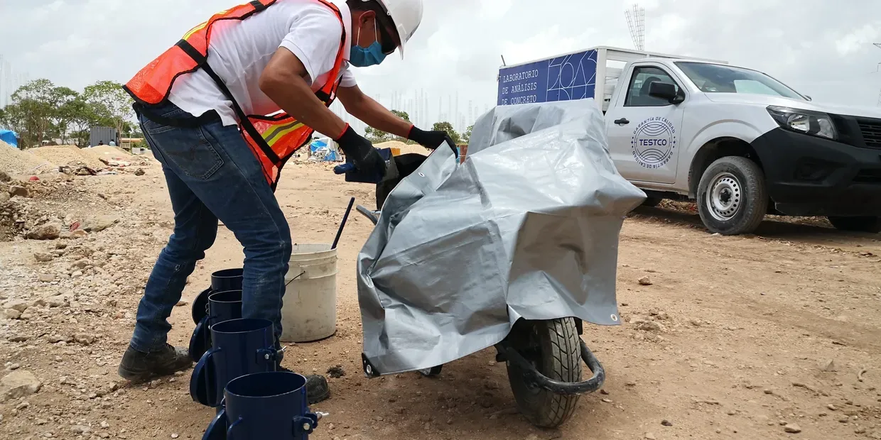 Técnico de TESTCO junto a moldes cilíndricos y una carretilla con concreto fresco en obra, con la unidad del laboratorio al fondo